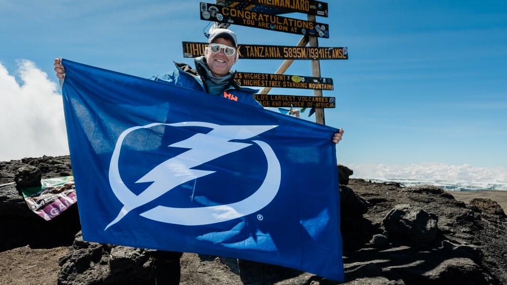 Preston Rudie stands at the summit of Mt Kilimanjaro holding a Tampa Bay Lightning flag. The summit signpost is behind. He is wearing wintry gear, including a jacket, hat, and protective glasses.