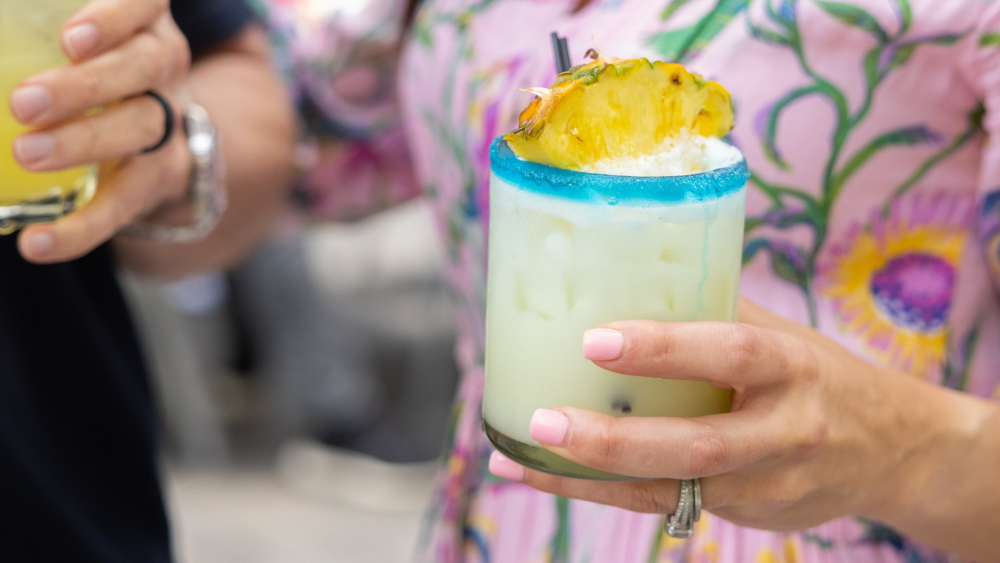 A woman in a floral dress holding a margarita with a blue rim and a pineapple