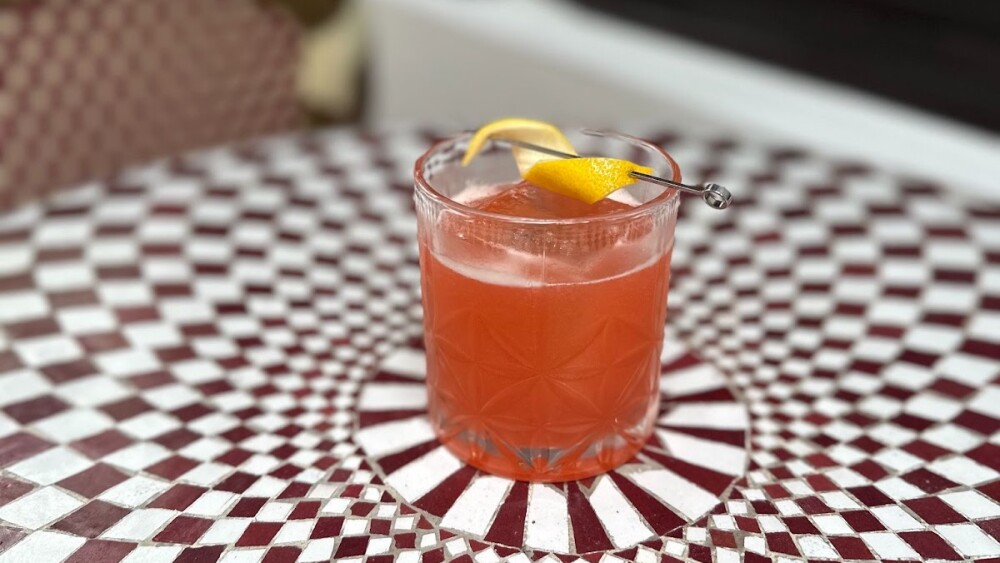 A peach-colored drink with a lemon peel garnish in a short whiskey glass sits on a brown and white tiled table.