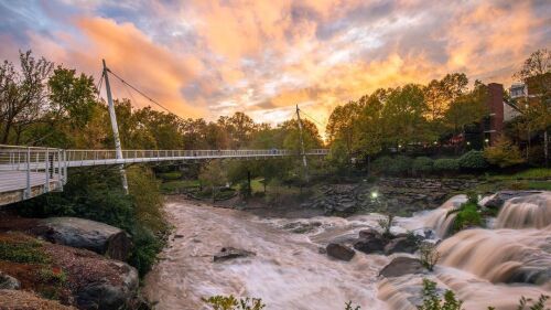 An orange and blue sunset over the bridge at Falls Park, with water rushing over the rocks. 