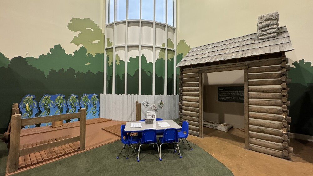 An area for children to explore the history of Charlotte with a faux farm area on the left, table and chairs in the center and a small log cabin on the right.