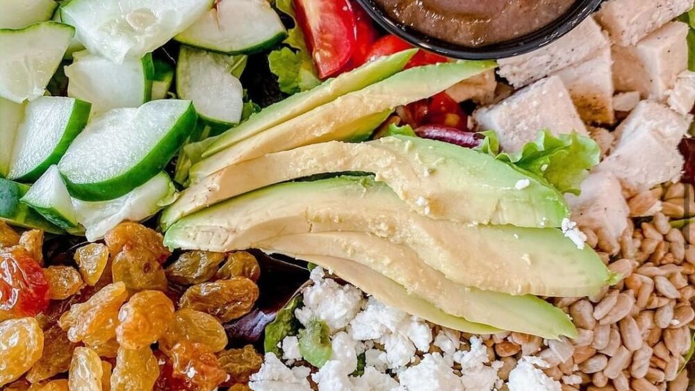 A salad with cheese, avocado, chicken, raisins, and nuts.