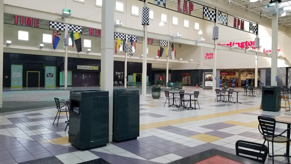 A large, open tiled area with multiple sets of tables and chairs sitting empty in front of a Burlington Coat Factory. Race Track decor is scattered throughout the space, including flags and signs that say words like "lap," "RPM," "time," and "race."