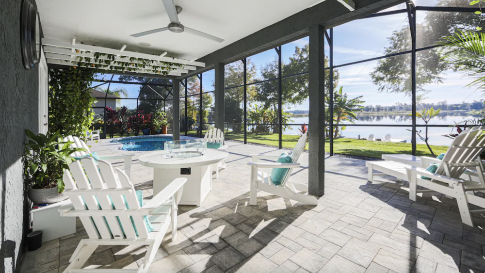 A beautiful screened-in patio overlooks a shining lake. Inside the patio, there are white beach chairs oriented around a small white table. Farther back, an in-ground swimming pool and hot tub occupy the back half of the patio.