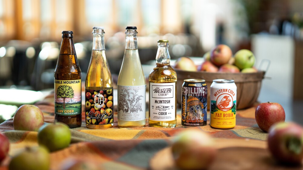 A variety of bottled and canned apple ciders sit on a table with apples