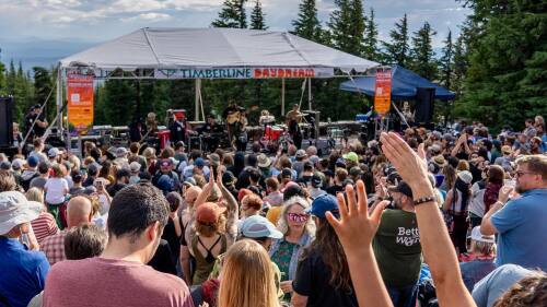 A crowd of people face a small outdoor stage in the mountains on a summer day.
