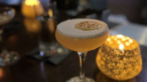 A yellow cocktail with a foamed top and dried lemon wedge floating on top next to a candle.