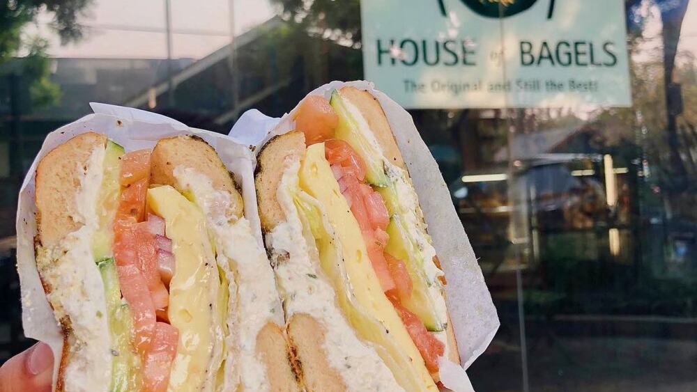 A hand holding a bagel with cream cheese, egg, tomatoes, and veggies in front of the "House of Bagels" sign.