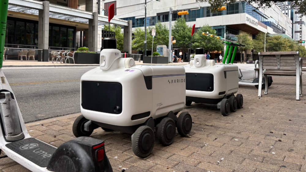Two cube-shaped little robots with cameras, sensors, and three wheels on each side are lined up on the Austin sidewalk, sandwiched by Lime scooters.