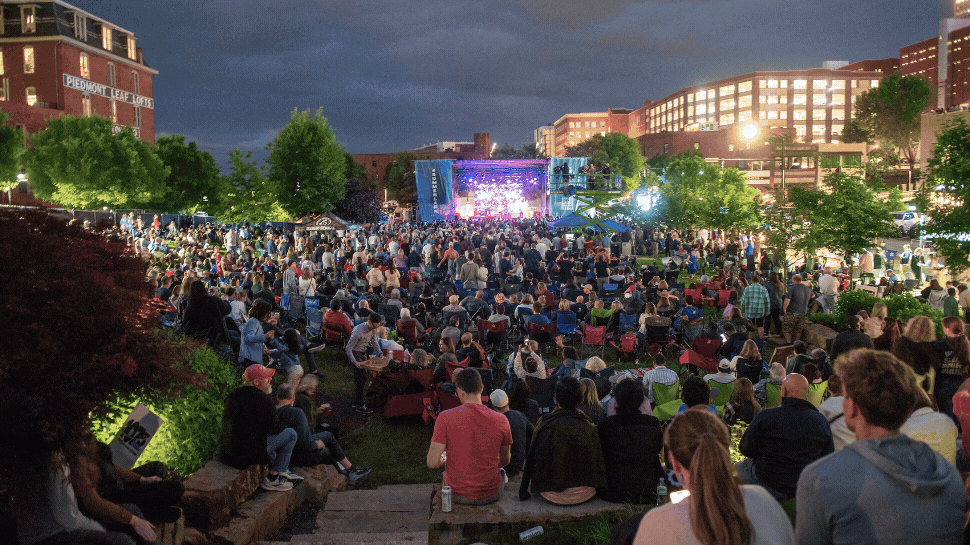 A crowd of people sits in a downtown park watching an outdoor concert.