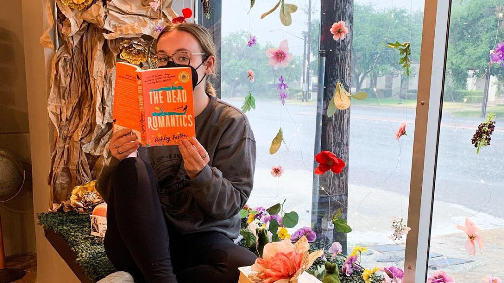 A person reading beside a stack of books in a window decorated with paper trees and flowers.