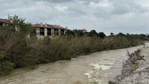 A muddy river flows swiftly under an overcast sky, with visible rapids and cloudy water. On the left side, several tan apartment buildings with red tile roofs sit behind a dense line of trees, some of which appear bent or damaged. The riverbank in the foreground is rocky and uneven.