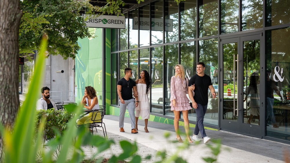 The exterior of Crisp and Green in Water Street Tampa. There are two couples walking in front of the glass storefront, arm in arm, and another couple sitting to the left at a metal table under a tree.