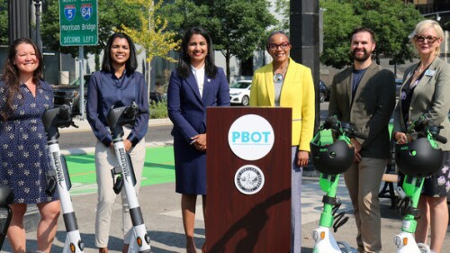 Leaders with the city of Portland, the Portland Bureau of Transportation, the scooter companies and community organizations stand next to a podium with four e-scooters propped in front of them on a city street.