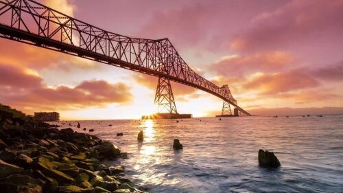 Looking up at a long truss bridge spanning a river before it meets the ocean. A stunning sunset has cast everything in varying shades of orange, pink, and blue.