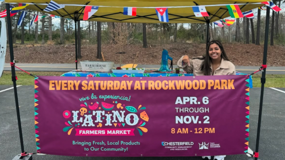 A vendor tent is set up, a banner of national flags handing from the top and a purple, orange, and blue sign for the Latino Farmers Market stretches across the front. A vendor behind the sign smiles and points down at the sign.