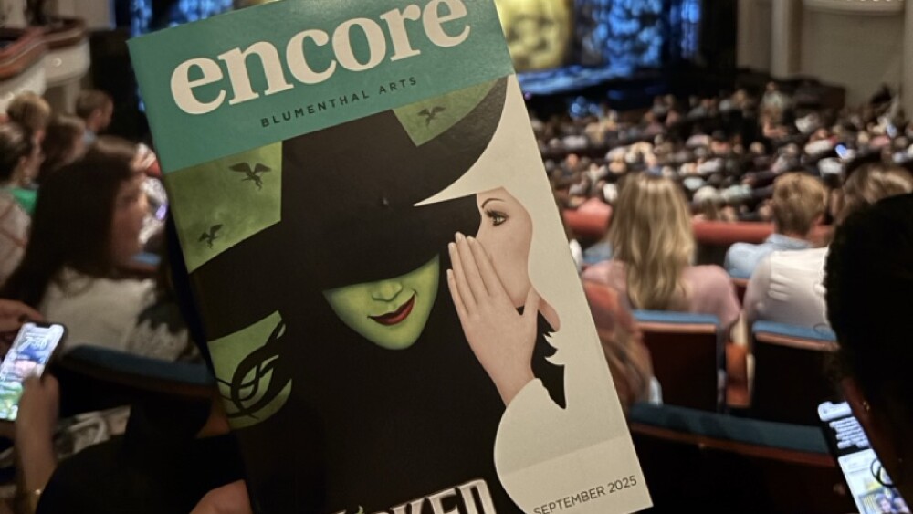 A playbill for the show "Wicked" with an audience and stage in the background.