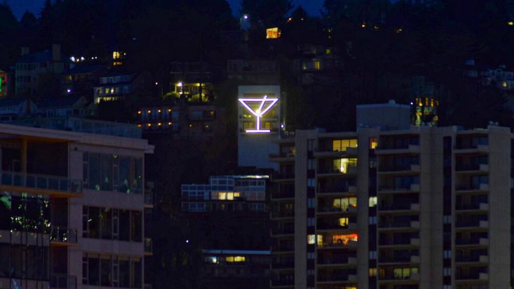 The illuminated outline of a martini glass lights up the night amidst residential buildings upon a hillside.