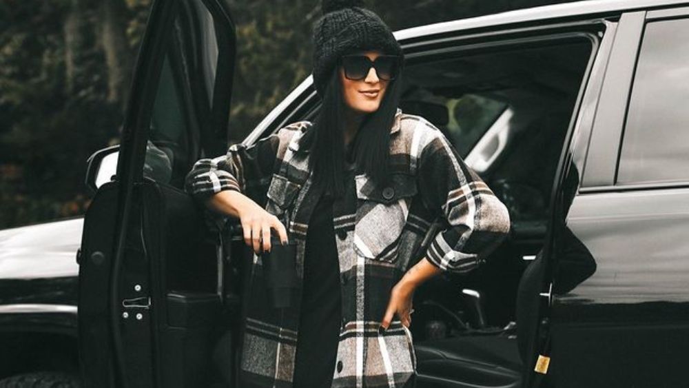 Influencer Ashley poses in a flannel shirt, beanie, and sunglasses in front of a black SUV in Austin, Texas.