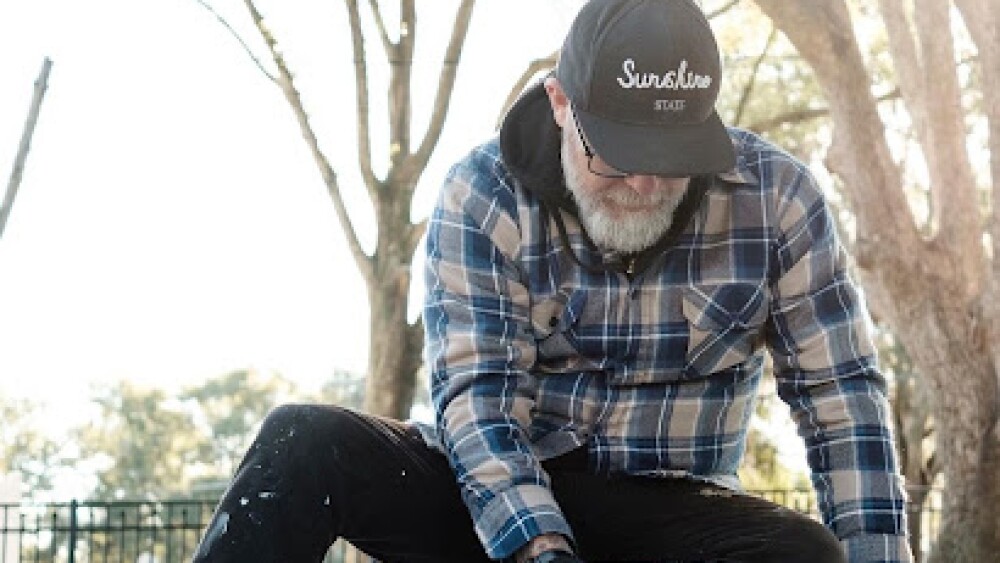 An artist wearing a baseball cap, plaid jacket, and skinny jeans crouches over a bucket of what appears to be paint.