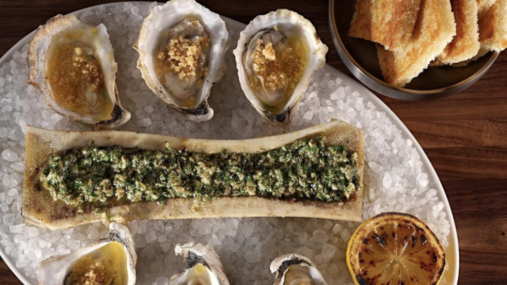 This elegant dish features six oysters topped with a savory garnish, flanking roasted bone marrow mixed with herbs. Served on rock salt with grilled lemon and crispy toast points on the side.