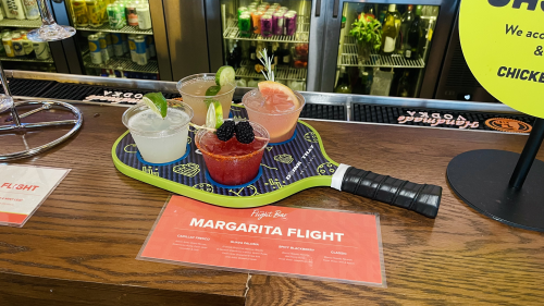Four cocktails garnished with fruit sit on a pickleball paddle on a bar. There's a sign in front of it that reads "margarita flight."