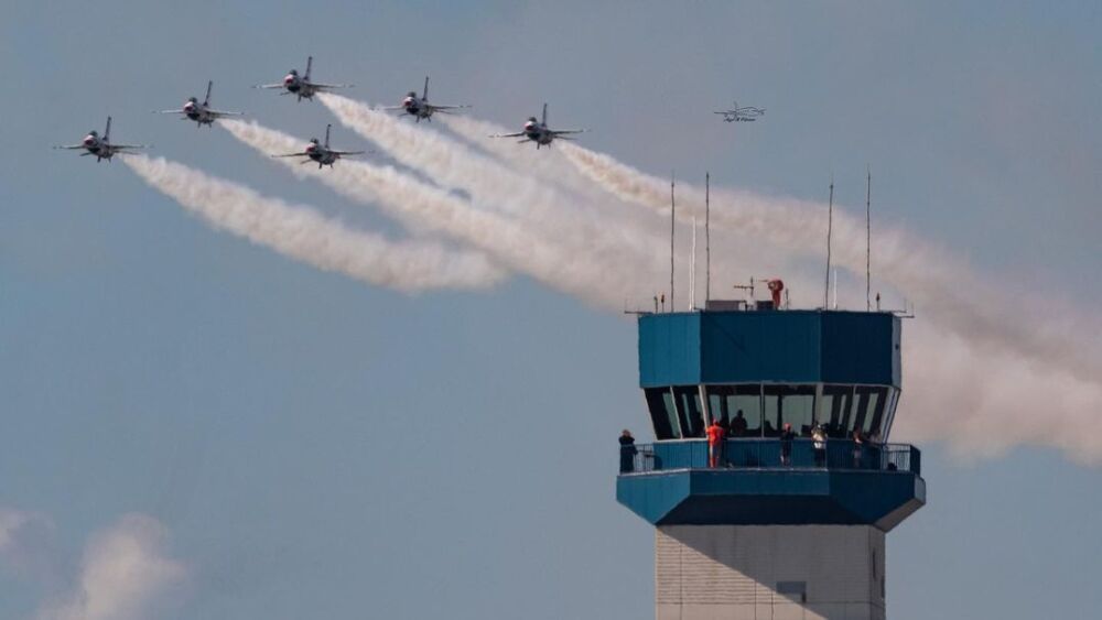 Six planes fly in a line formation over a control tower at SUN 'n FUN. Each plane has a cloud of vapor trails following it.