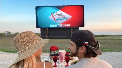 A couple has their backs to the camera and they are looking at one another. In front of them is a projector screen outdoors and a table with popcorn and drinks on it.