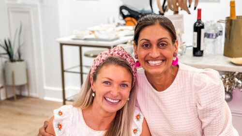 Two women smile brightly at a social gathering. One, wearing a name tag labeled "Amanda," holds a Mahjong tile rack, suggesting they’re playing the game. Both wear pink-themed outfits and accessories. Behind them, a table with snacks and drinks, colorful artwork, and headbands creates a cheerful, creative atmosphere.
