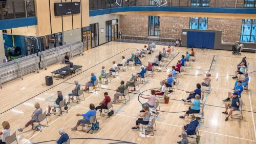 Dozens of seniors take part in a chair yoga class in a gymnasium.