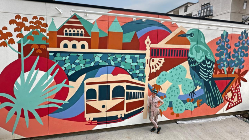 A mural depicting the Dallas County Courthouse, a mockingbird, cacti, and a trolley is painted along the side of a building using a dusty rose + teal color palette.