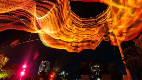 A moving wire sculpture is lit up orange and moves above the city skyline.