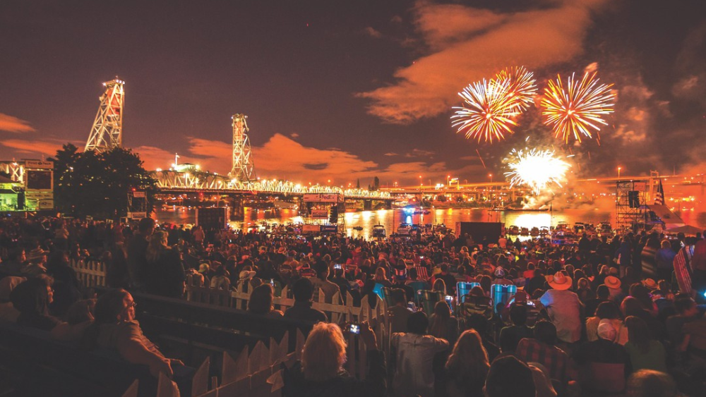 Fireworks explode above a packed crowd at Portland's Waterfront Blues Festival, illuminating the nearby Hawthorne Bridge and soundstage in an orange glow.