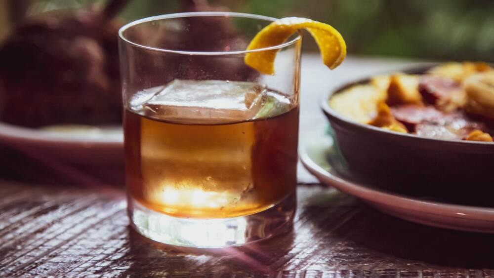 A glass of whiskey with a lemon peel garnish and a large ice cube.