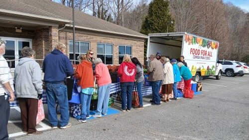 A diverse group of people gathers around tables with food items outside a building. A truck labeled "Food for All" is parked nearby.
