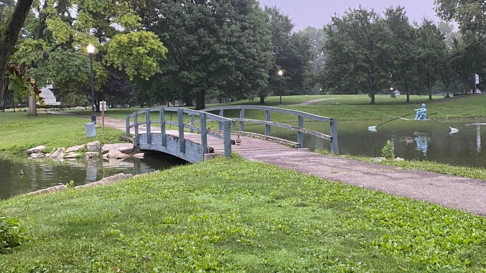 A bridge crossing a pond with a statue of a rower to the right.