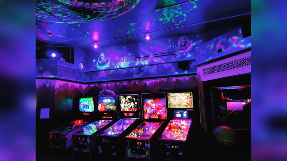 A dark room with five brightly colored pinball machines and cosmic glow-in-the-dark art on the walls at Space Room Lounge in Portland, Oregon.