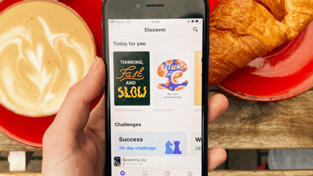 A hand holding a phone open to the Headway app, in front of a coffee and croissant.