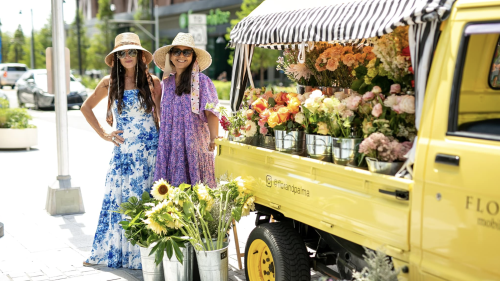 Two women in sundresses standing in front of a yellow truck full of flowers.