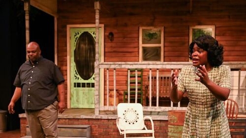 2 actors stand in front of a set that features a brown wooden house with steps leading up to a green front door, a white lawn chair and barrel stand in the front yard of the set