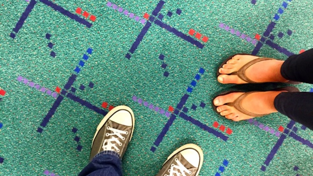 An aerial view looking down at two peoples' feet as they stand on teal-colored carpet with multi-colored designs.