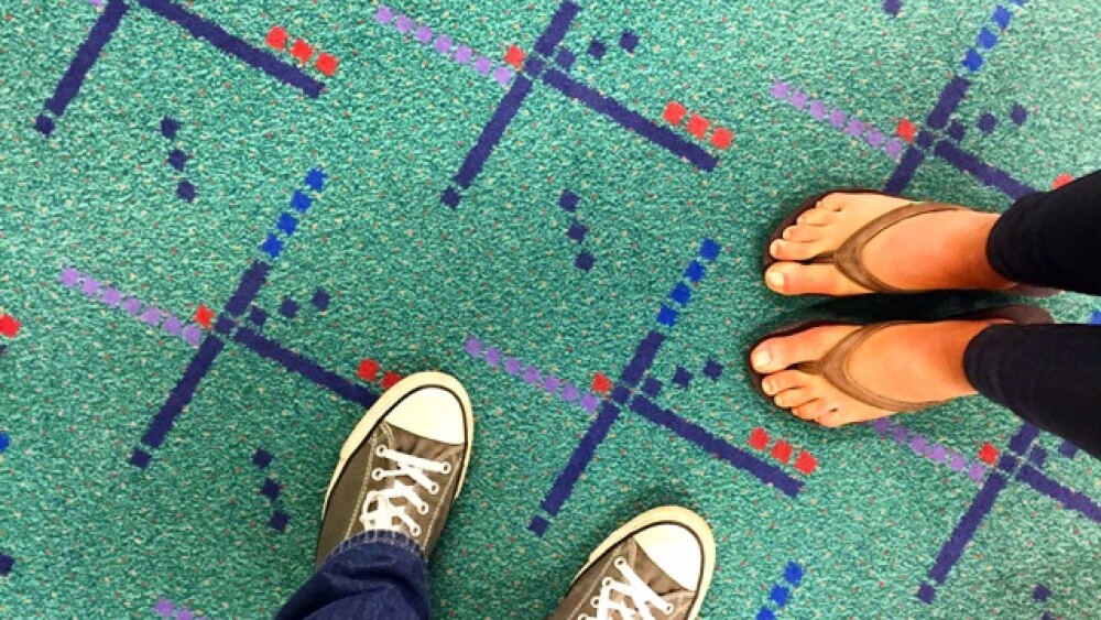 An aerial view looking down at two peoples' feet as they stand on teal-colored carpet with multi-colored designs. 