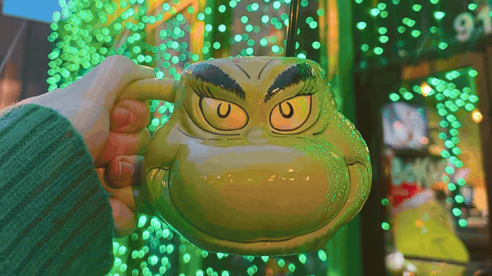 A mug in the shape of the Grinch's head in front of green string lights.
