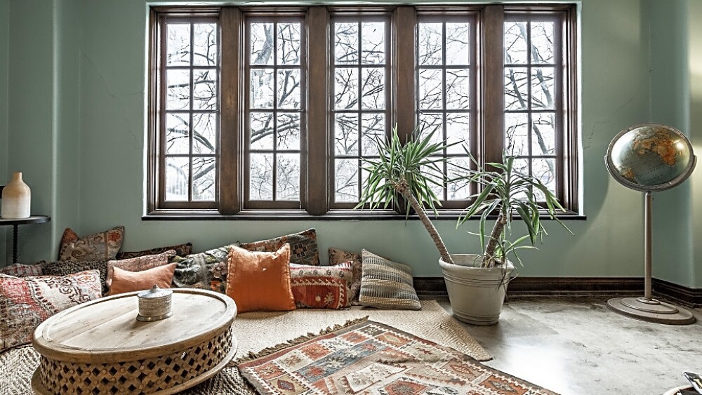 A cozy room with large windows and lots of pillows on the floor with a table and rug