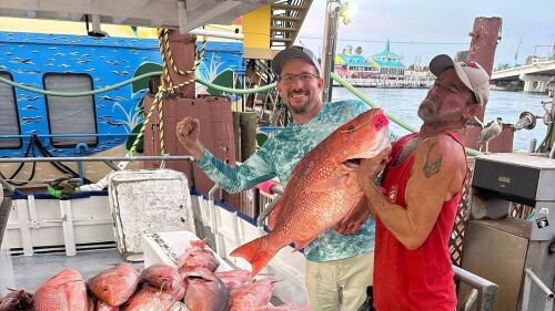 Two men stand behind a pile of fish and one of the men holds one fish up on a Hubbard's Marina boat with other boats and buildings in the background.