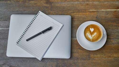 A top-down view of a laptop sitting on a table with an open notebook and pen sitting on top of it. Next to the laptop is a latte in a mug atop a white saucer. The latte has a foam heart on top.