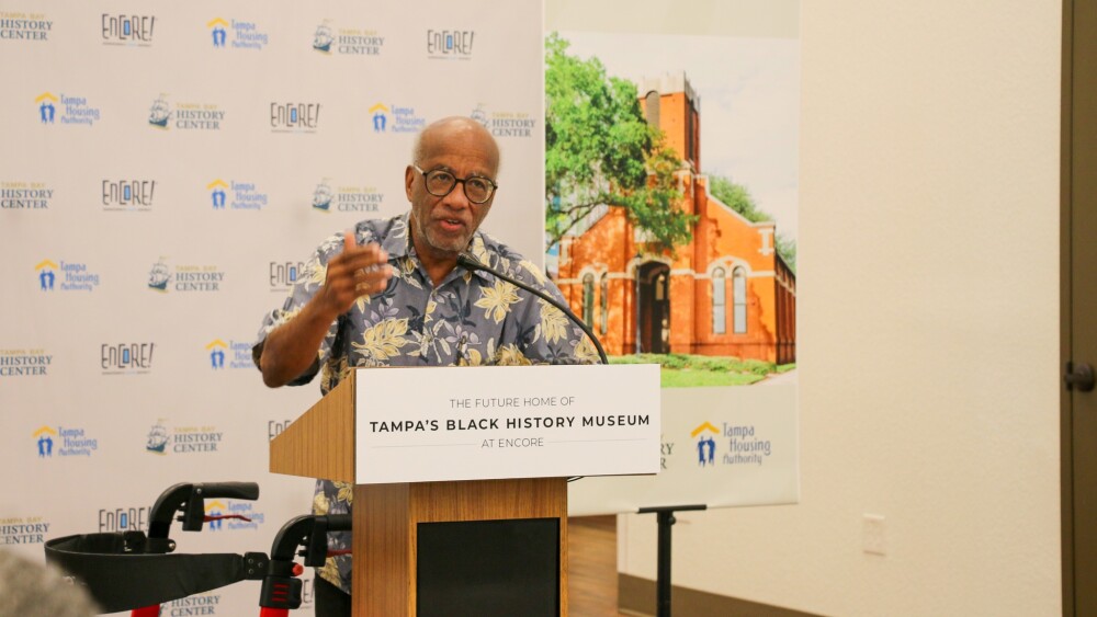 A speaker at the podium during the official announcement of Tampa's first ever Black History Museum. The man is wearing a blue and yellow flowery shirt, and a picture of the church is behind him.