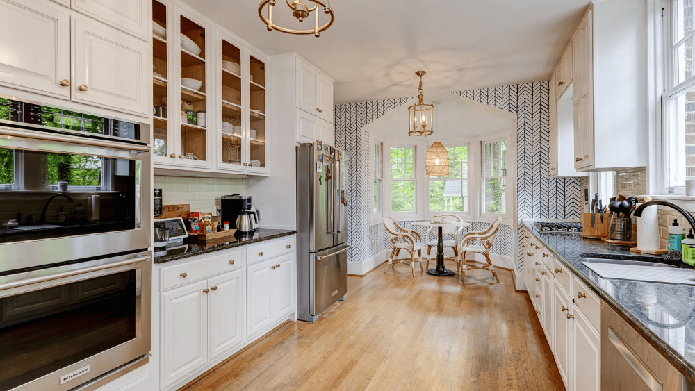 A kitchen with a dining nook complete with a wicker light fixture, blue wallpaper, and a small table and chair set overlooking a bay window.