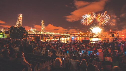 Fireworks explode over a river as crowds of people look on. An orange glow lights up the city and a bridge.