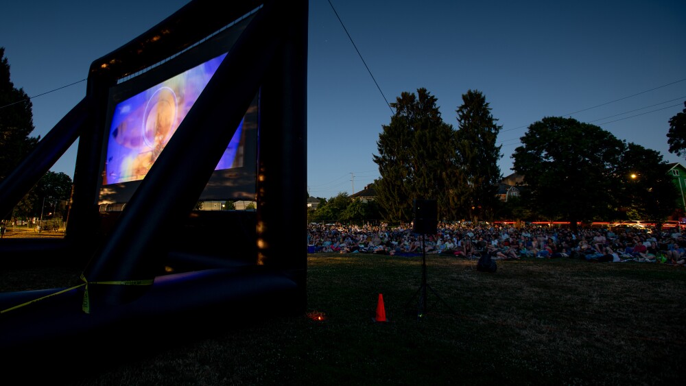 Colorful images illuminate a movie projector screen held upright by a large frame at dusk as a crowd of people seated on a grassy park field look on.